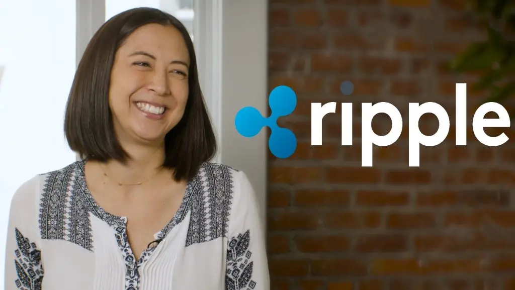 Monica Long, President of Ripple, smiling in front of a brick wall and the Ripple logo